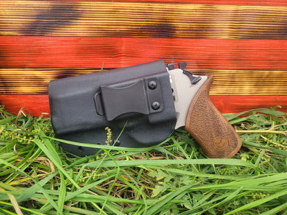 A firearm in a black holster rests on green grass with a colorful backdrop of red and gold stripes.
